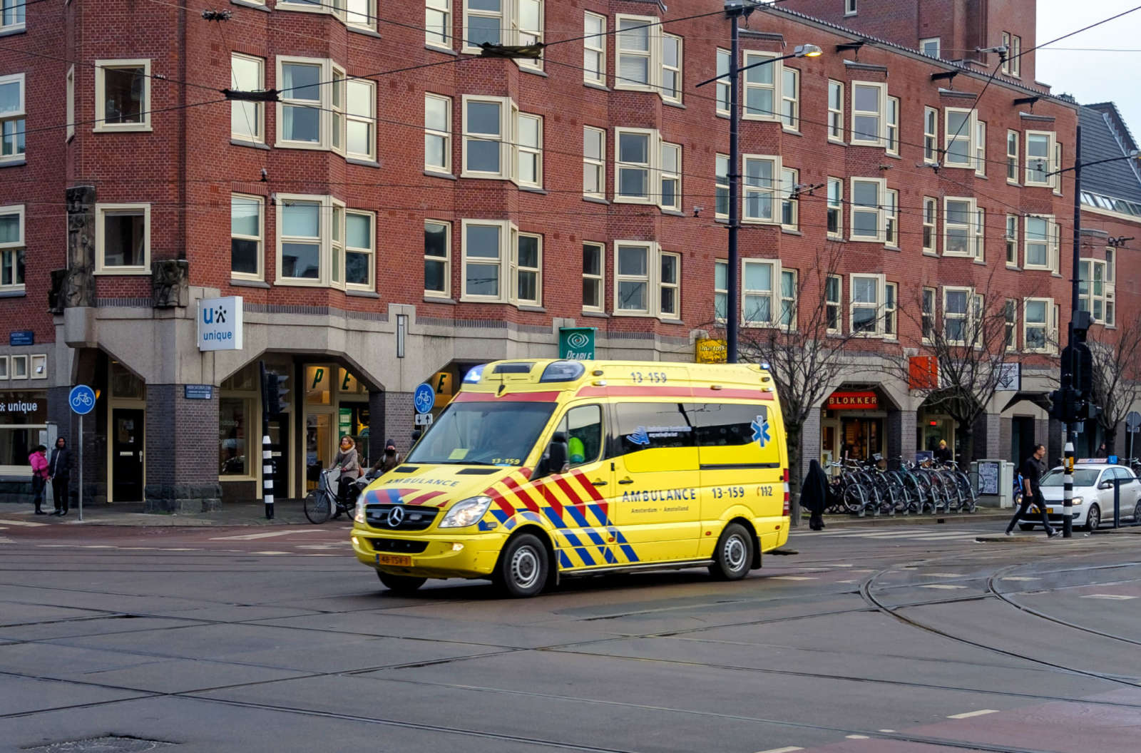 A Dutch ambulance with flashing lights crossing a city intersection with tram tracks, past cyclists and pedestrians.