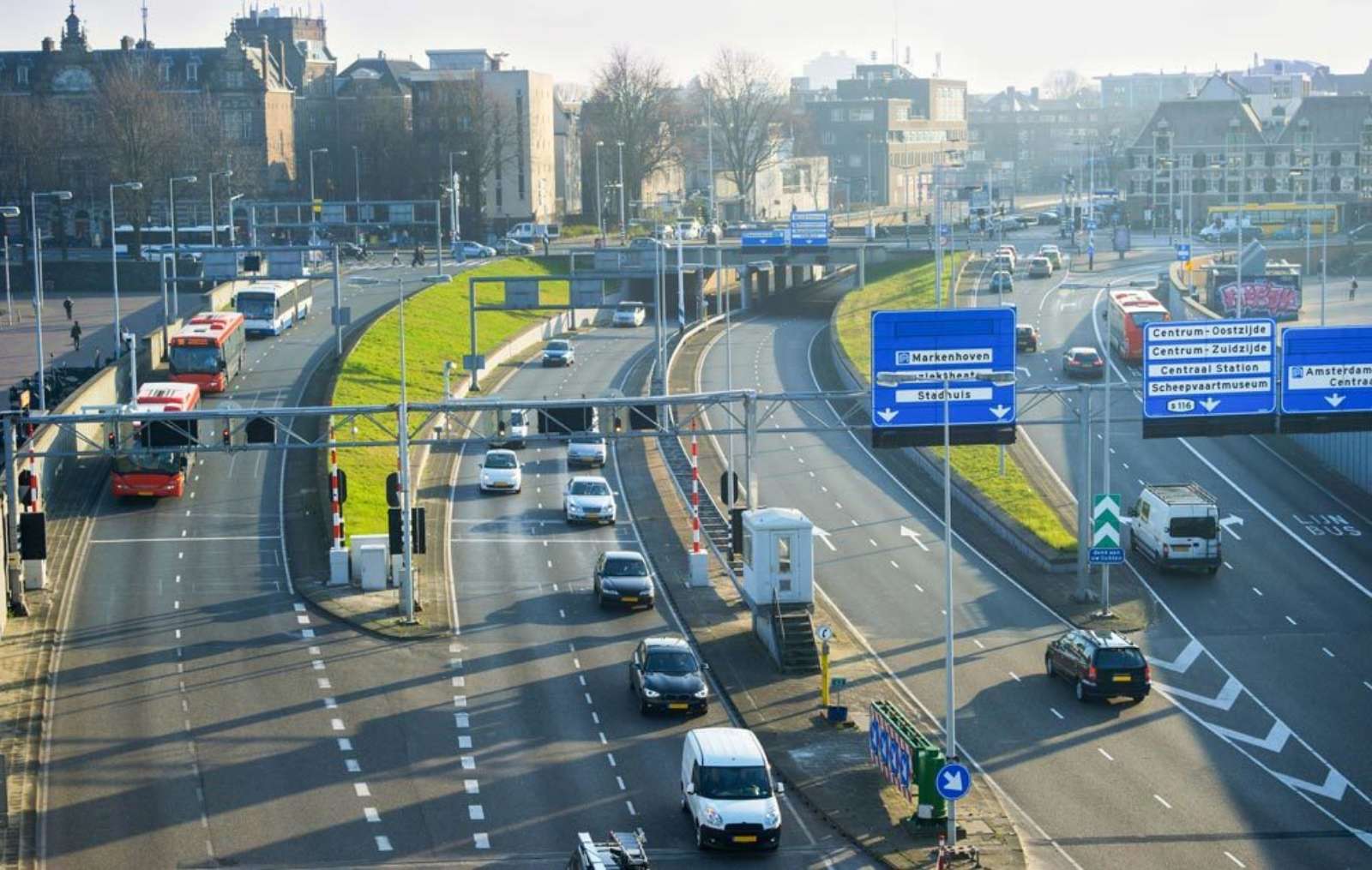 Multi-lane urban road with buses on a dedicated lane, tunnel entrances and blue overhead direction signs towards Amsterdam city centre.