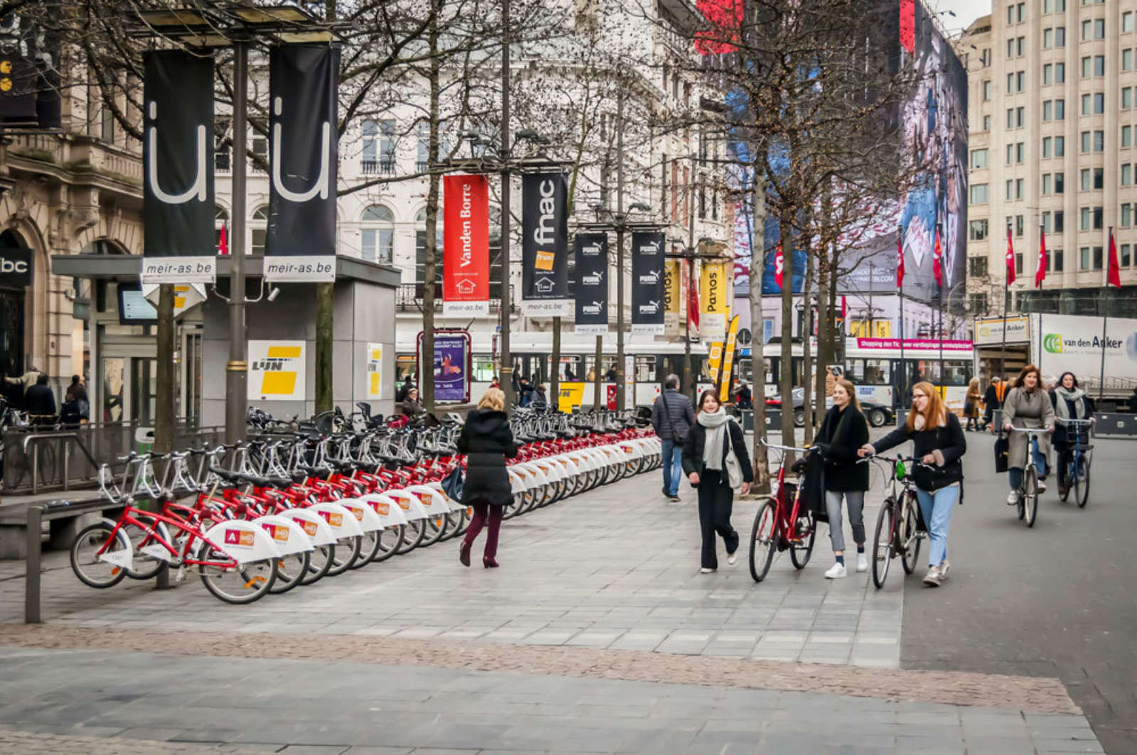A long row of red and white shared bikes on the Meir in Antwerp, with pedestrians and cyclists passing by.