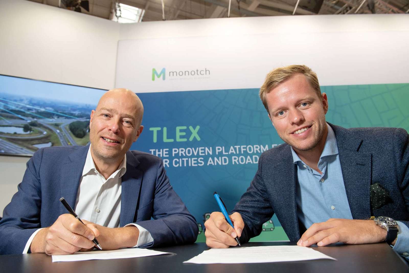 Menno Malta of Monotch and Jorn de Vries of Be-Mobile signing the partnership agreement at a trade fair stand, in front of a TLEX banner.