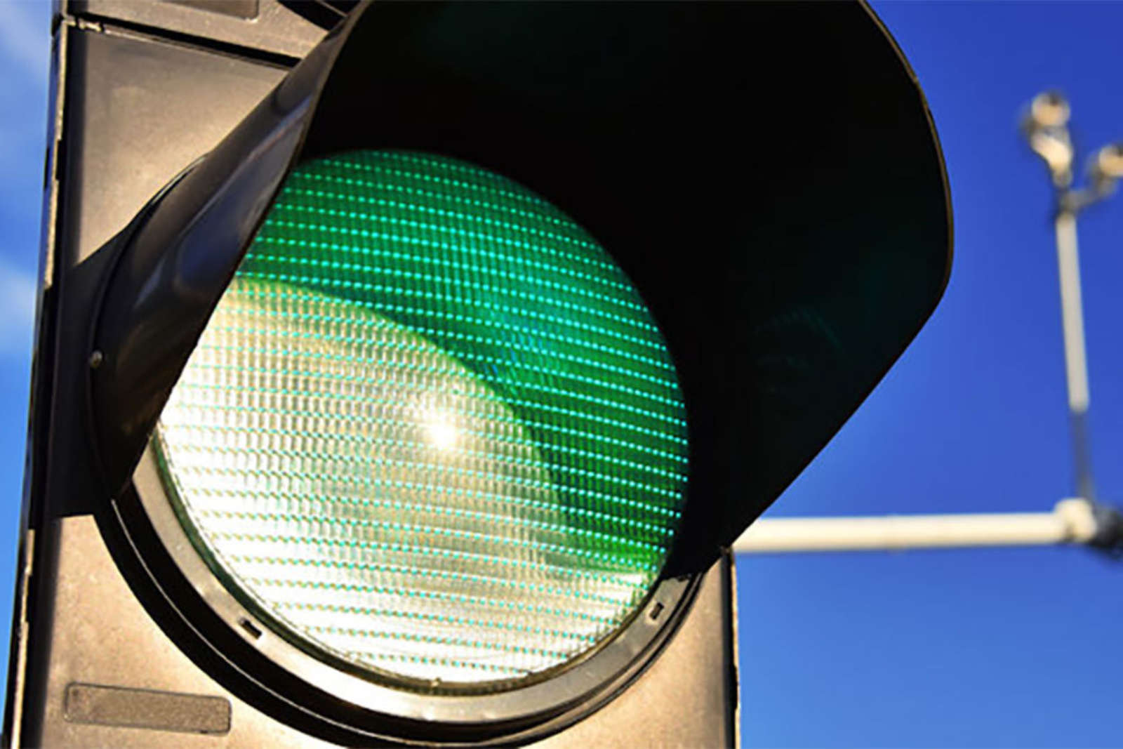 Close-up of a green traffic light against a blue sky, with a camera mast in the background.